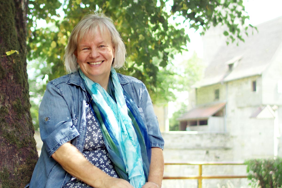 Märchen­pädagogin & Naturtherapeutin Elke Fischer-Wagemann sitzt lächelnd vor einer Weide und schaut in die Kamera, während sie über ihre Angebote und die Zusammenarbeit mit sich spricht.