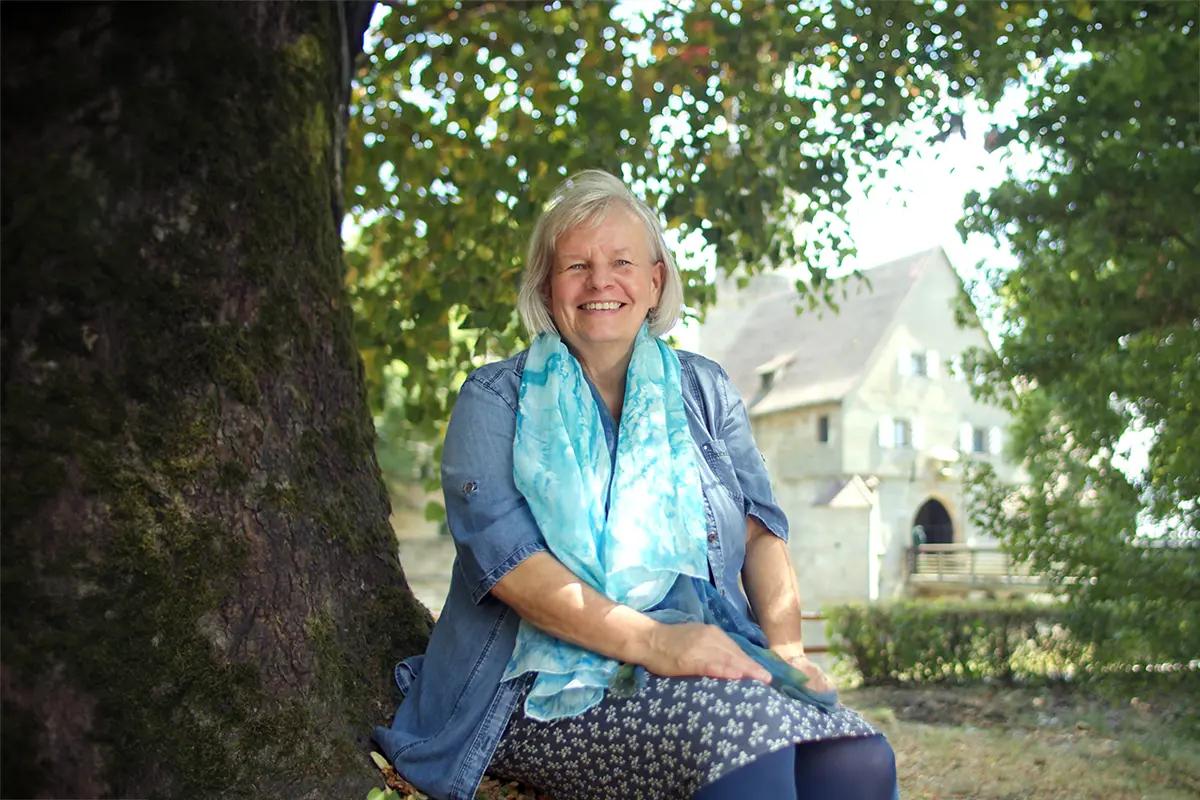 Naturtherapeutin Elke Fischer-Wagemann sitzt unter einem Baum und lächelt offen in die Kamera. Im Hintergrund sieht man eine alte Burg.